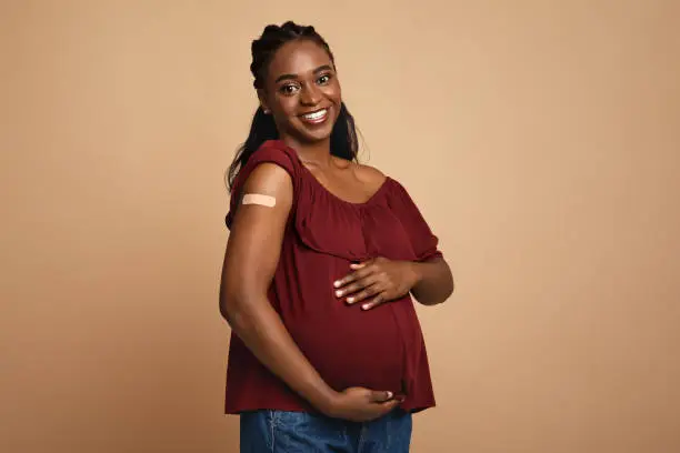Safety vaccination during pregnancy cocnept. Cheerful pregnant young black woman got vaccinated against COVID-19, showing her arm with band on and hugging her big tummy, expecting baby, copy space