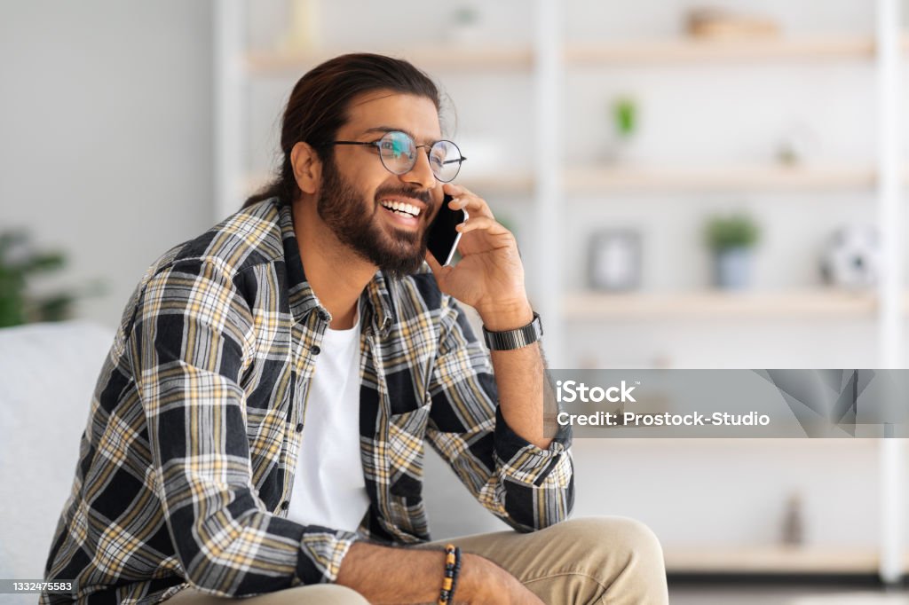 Cheerful guy relaxing on couch at home, having phone talk Cheerful arab guy with long hair and glasses relaxing on couch at home, having phone talk with friends, looking at copy space and smiling, wearing casual outfit, resting at weekend Using Phone Stock Photo