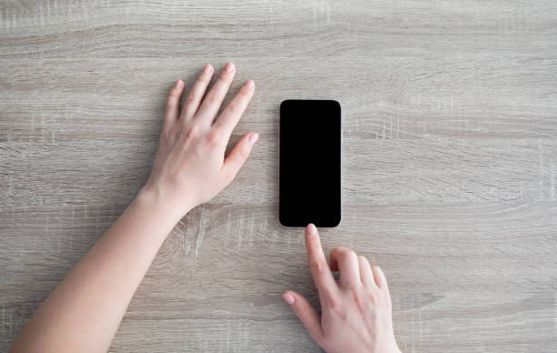 istock Female hands touching modern smartphone with blank black screen for mockup on wooden table background, top view 1332476480