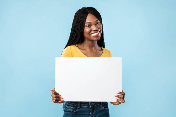 positive afroamerikanerin zeigt leeren karton, blauer hintergrund - nun sign holding women stock-fotos und bilder