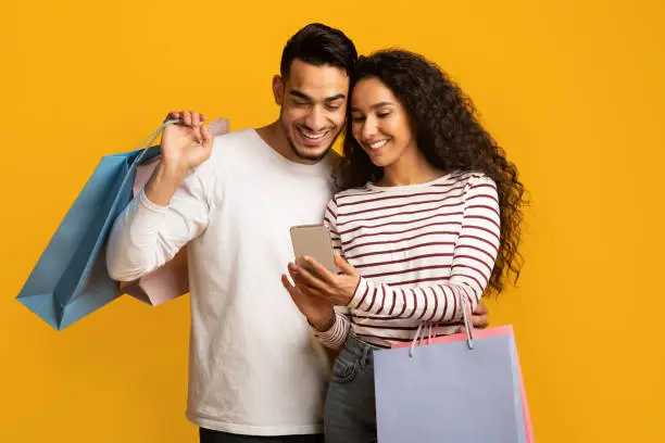 Discount App. Cheerful Arab Couple Holding Smartphone And Colorful Paper Shopping Bags, Middle Eastern Spouses Browsing Mobile Application With Coupons, Promo Codes, and Deals Over Yellow Background