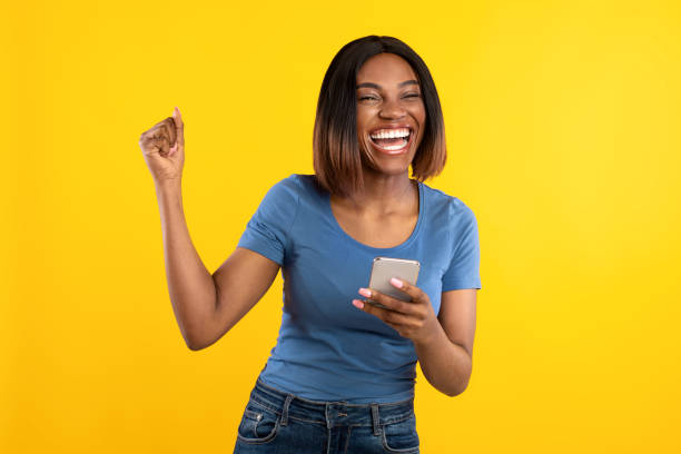 happy black lady sosteniendo el teléfono gesticulando sí sobre fondo amarillo - eufórico fotografías e imágenes de stock