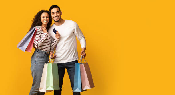 Easy Shopping. Happy Arab Couple Holding Credit Card And Bright Shopper Bags Easy Shopping. Happy Arab Couple Holding Credit Card And Bright Shopper Bags, Cheerful Middle Eastern Spouses Carrying Purchases, Posing Over Yellow Studio Background, Copy Space, Panorama shopping photos stock pictures, royalty-free photos & images