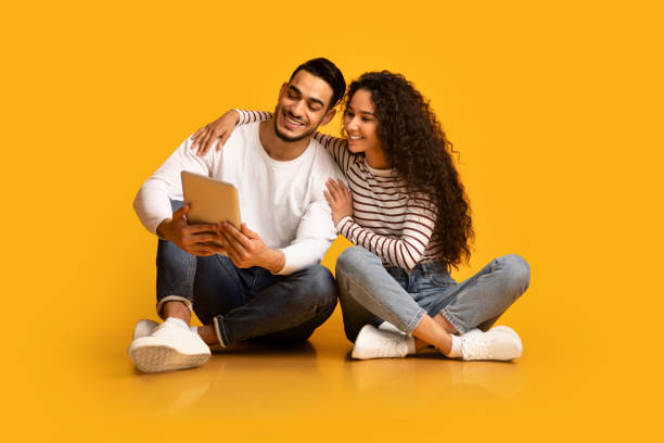 felice giovane coppia mediorientale che usa tablet digitale mentre è seduta su sfondo giallo - floor sitting foto e immagini stock