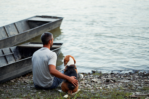 A young man is sitting by the river next to the docked rowboats with his dog.