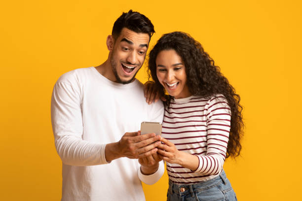 cool app. pareja árabe emocionada mirando la pantalla del teléfono inteligente en las manos - couple young women casual excitement fotografías e imágenes de stock