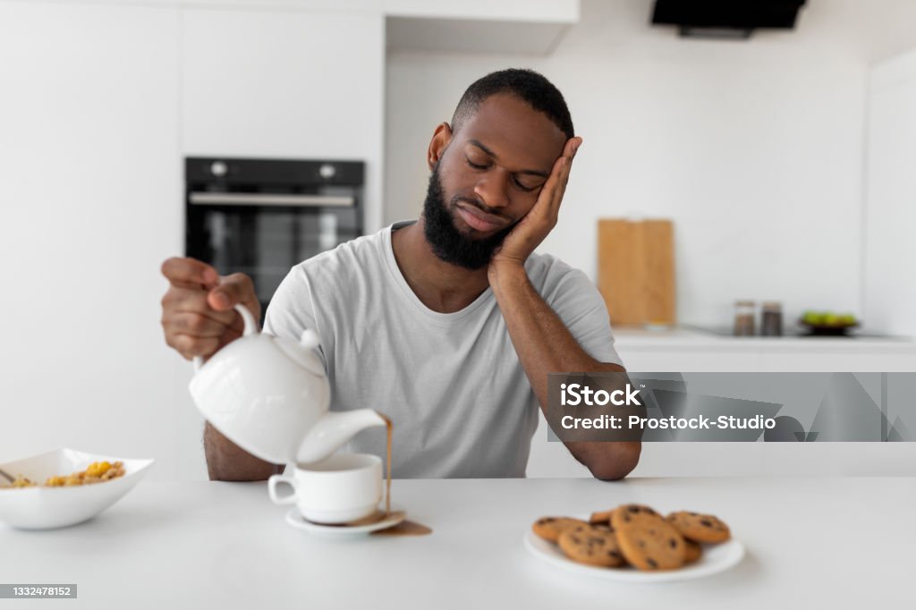 Black man pouring coffee away from cup spilling hot drink Lack Of Sleep Concept. Portrait of tired young African American man sleeping while sitting at dining table in kitchen, holding kettle pouring coffee away from cup spilling hot drink on desk Sleeping Stock Photo