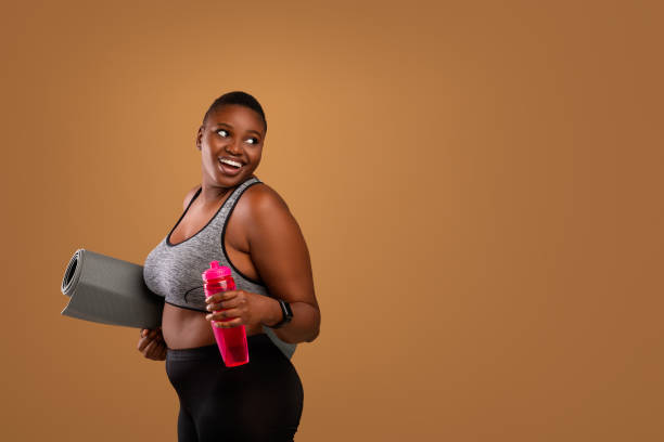 Young Black Woman Posing With Mat And Water At Studio Body Positivity And Sport. Smiling Curvy Black Woman In Sportswear Holding Fitness Mat And Bottle With Water, Looking Back At Free Copy Space For Advertisement, Standing Isolated On Brown Background huge black woman pictures stock pictures, royalty-free photos & images