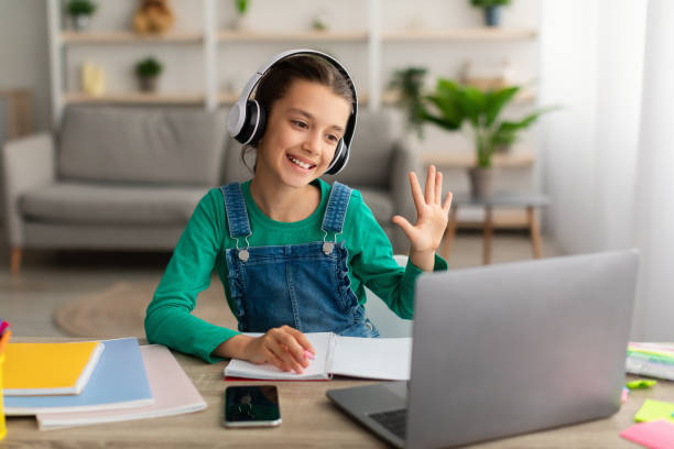 istock Girl sitting at table, using laptop, waving to webcam 1332478256
