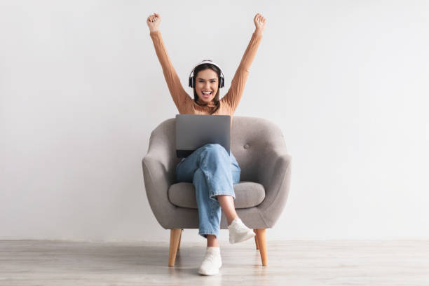 mujer joven emocionada con auriculares sentada en el sillón, celebrando el triunfo en línea, gran oferta o éxito empresarial - full length audio fotografías e imágenes de stock