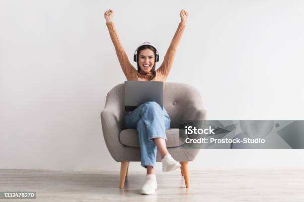 Excited Young Woman In Headphones Sitting In Armchair Celebrating Online Win Great Deal Or Business Success Stock Photo - Download Image Now