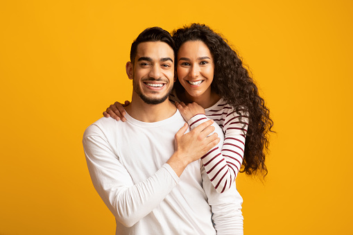 Happy Arab Spouses. Closeup portrait of cheerful middle eastern couple hugging and smiling at camera, joyful millennial lovers standing over yellow background, posing together in studio, copy space