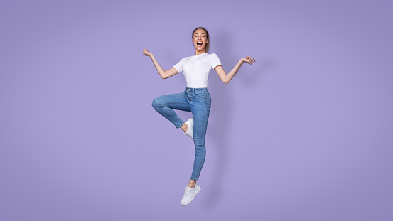 Cheerful Blonde Millennial Female Jumping Smiling To Camera Over Purple Background. Studio Shot Of Carefree Young Woman Posing In Mid-Air. Meditation And Relaxation Concept. Full Length, Panorama