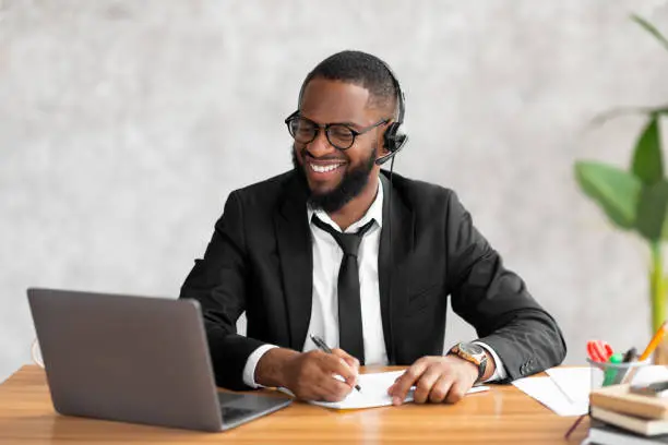 Smiling African business man wearing suit, eyeglasses and headset working on laptop from home office. Black man writing in notebook taking notes and using computer, sitting at desk watching webinar