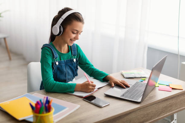 istock Girl sitting at table, using laptop, writing in textbook 1332478659
