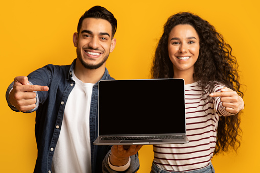 Great Website. Smiling middle eastern couple pointing at laptop with black screen for mockup, demonstrating copy space for your web design and advertisement, standing isolated over yellow background