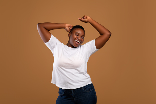 Taking Break. Overjoyed millennial plus-size black woman wearing white t-shirt dancing with closed eyes, having fun moving and listening to music, relaxing isolated over dark brown studio background