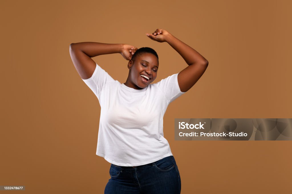 Portrait of casual young black woman dancing, brown wall Taking Break. Overjoyed millennial plus-size black woman wearing white t-shirt dancing with closed eyes, having fun moving and listening to music, relaxing isolated over dark brown studio background Women Stock Photo