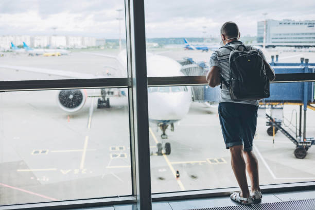 istock Man in protective medical face mask with backpack standing at empty airport waiting for departure. Male passenger in T-shit looking out window at airplane. Travel during coronavirus virus pandemic 1332480824