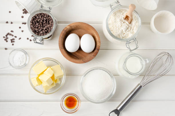 istock Ingredients for making muffins with chocolate chips. Eggs, butter, sugar, flour, vanilla, chocolate chips on a white wooden background. Recipe step by step. Top view. 1332483573