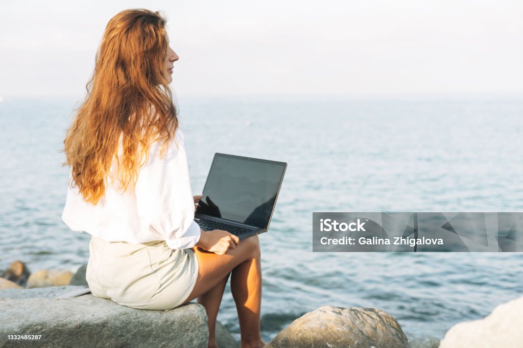 Young carefree beautiful freelance businesswoman with long hair in a white shirt working at laptop on seashore on sunrise Young carefree beautiful freelance businesswoman with long hair in a white shirt working at laptop on the seashore on sunrise 35-39 Years Stock Photo