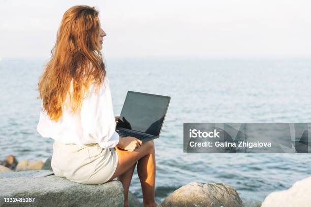 Young Carefree Beautiful Freelance Businesswoman With Long Hair In A White Shirt Working At Laptop On Seashore On Sunrise Stock Photo - Download Image Now