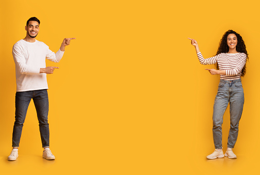 Check This. Happy arab couple pointing at copy space in the middle of yellow background, young middle eastern man and woman recommending something, showing free place for your design or advertisement