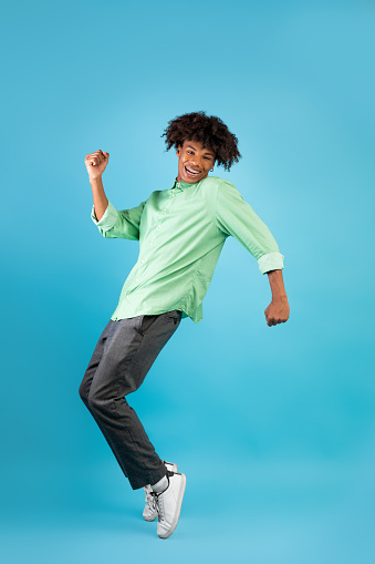 Joyful african american teenage guy standing on tiptoes, dancing, fooling and having fun over blue background, studio shot, copy space, crop