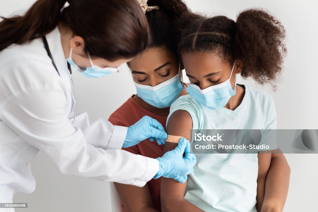 African american family making vaccination at hospital, wearing face masks African american family mother and cute daughter making vaccination at clinic, wearing protective face masks. Doctor putting band on kid shoulder after injection against coronavirus, closeup Vaccination Stock Photo