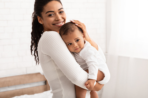 Mommy And Baby Son. African American Mother Posing With Little Infant Boy Holding Her Lovely Son In Hands Standing In Bedroom At Home, Smiling To Camera. Mom's Lifestyle, Child Care