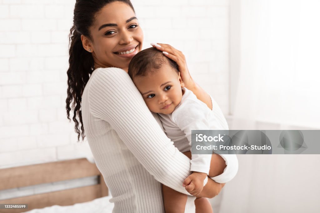 African American Mother Holding Her Baby Son Standing In Bedroom Mommy And Baby Son. African American Mother Posing With Little Infant Boy Holding Her Lovely Son In Hands Standing In Bedroom At Home, Smiling To Camera. Mom's Lifestyle, Child Care Mother Stock Photo