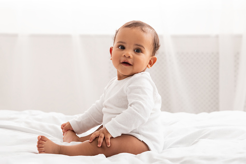 Infancy. Adorable African American Infant Baby Sitting In Bed Smiling To Camera At Home. Cute Toddler Boy Posing Wearing White Longsleeve Coverall In Bedroom Indoor. Babies Fashion And Clothes