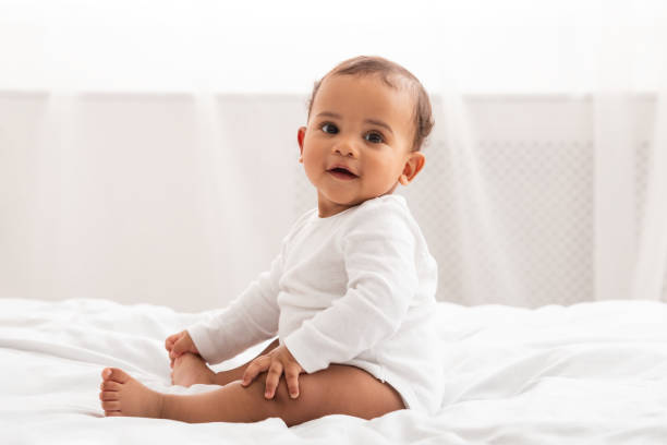 African American Infant Baby Sitting In Bed Smiling At Home Infancy. Adorable African American Infant Baby Sitting In Bed Smiling To Camera At Home. Cute Toddler Boy Posing Wearing White Longsleeve Coverall In Bedroom Indoor. Babies Fashion And Clothes nanny photos stock pictures, royalty-free photos & images