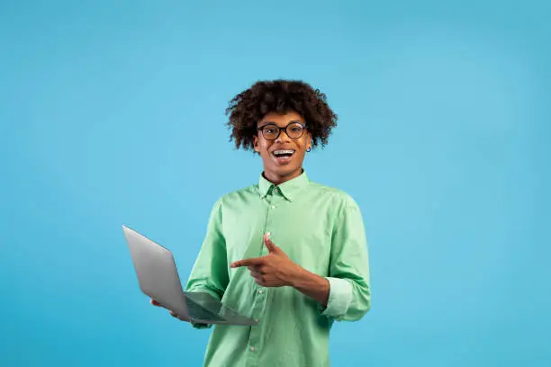 Online education concept. Happy african american teenage student using laptop computer for e-learning, pointing finger at pc, standing over blue studio background