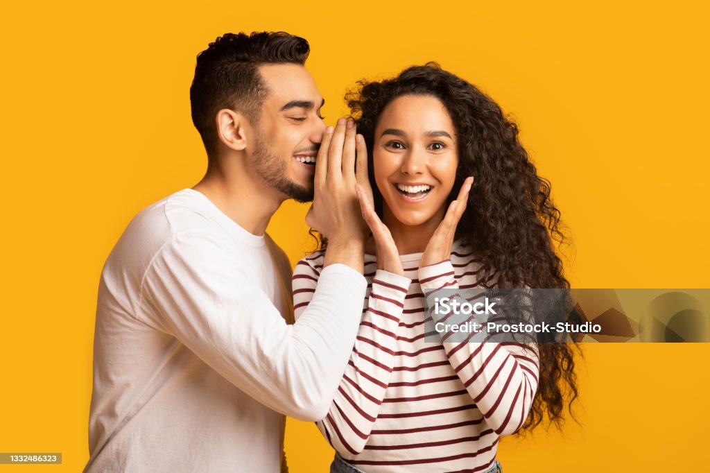Big Secret. Young Arab Man Sharing News With His Excited Girlfriend Big Secret. Young Arab Man Sharing News With His Excited Girlfriend, Surprised Middle Eastern Woman Raising Hands In Amazement While Standing Together Over Yellow Studio Background, Free Space Whispering Stock Photo