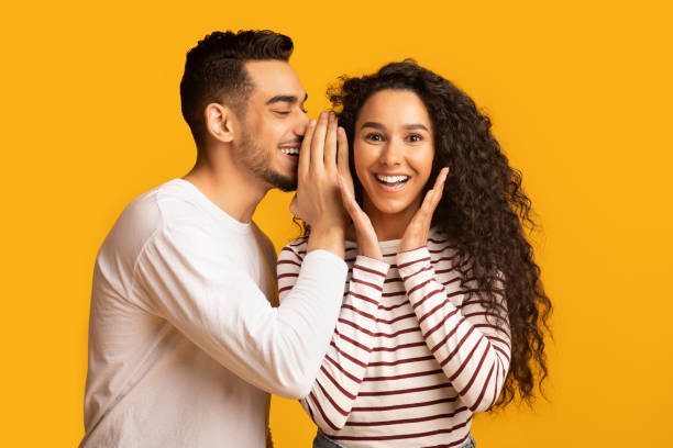 Big Secret. Young Arab Man Sharing News With His Excited Girlfriend Big Secret. Young Arab Man Sharing News With His Excited Girlfriend, Surprised Middle Eastern Woman Raising Hands In Amazement While Standing Together Over Yellow Studio Background, Free Space gossip stock pictures, royalty-free photos & images