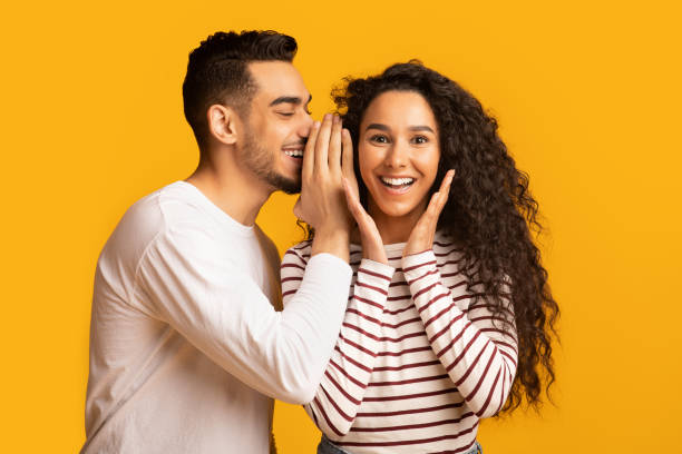 Big Secret. Young Arab Man Sharing News With His Excited Girlfriend Big Secret. Young Arab Man Sharing News With His Excited Girlfriend, Surprised Middle Eastern Woman Raising Hands In Amazement While Standing Together Over Yellow Studio Background, Free Space whispering stock pictures, royalty-free photos & images