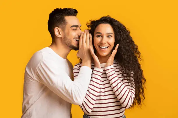 Big Secret. Young Arab Man Sharing News With His Excited Girlfriend, Surprised Middle Eastern Woman Raising Hands In Amazement While Standing Together Over Yellow Studio Background, Free Space