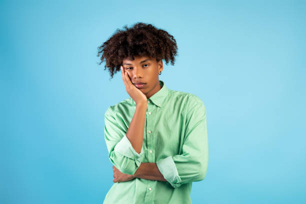 portrait d’un adolescent noir qui s’ennuie, qui s’est penché sur sa main, qui passe une journée terne sur un fond de studio bleu - dull colors photos et images de collection