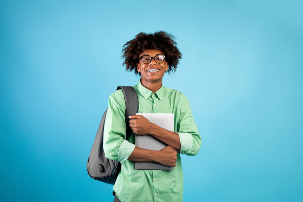 istock Student's lifestyle concept. Happy african american guy holding laptop hugging computer, standing on blue background 1332486417