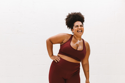 Plus size woman smiling with closed eyes with one hand on her waist. Pretty female in sportswear relaxing after exercise on white background.