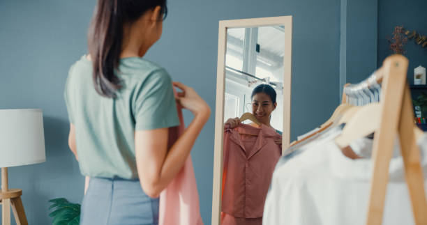 家のリビングルームで鏡で自分自身を見て服ラックドレッシングに服を選ぶ美しい魅力的なアジアの女性。 - garment ストックフォトと画像