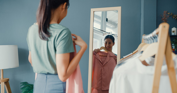 Beautiful attractive Asia lady choosing clothes on clothes rack dressing looking herself in mirror in living room at house. Girl think what to wear casual shirt. Lifestyle women relax at home concept.