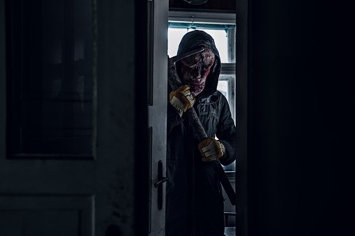 Scary man wearing a terrifying mask with a pick-axe, standing behind the door at night. Scene from a horror story, man in disguise standing next to the door in a dark spooky house.