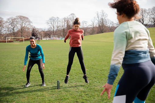 A diverse group of women exercising, jumping in the air doing burpees in a public park surrounded by nature. They are working out in their community to get exercise and spend quality time together.