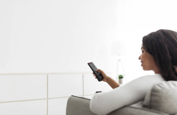 istock Young black lady holding and pointing remote control on white empty wall, mockup for your design or advertisement 1332488041