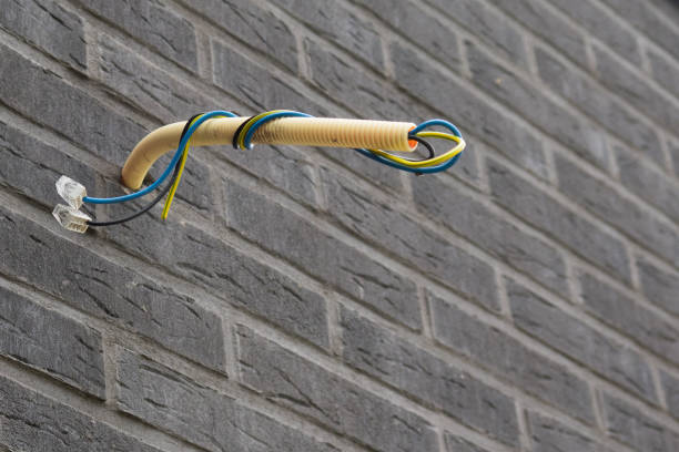 Uit een grijze bakstenen muur steekt een gele flexibele elektriciteitsbuis met daarin drie draden: een blauwe, een zwarte en een geelgroene. Aan de uiteinden van de zwarte en blauwe draad zitten transparante lasdoppen.