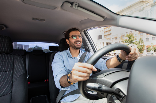 Joyful indian man driving car, shot from dashboard, going on trip during summer vacation, copy space. Happy middle-eastern guy in casual outfit and glasses driving his brand new nice car