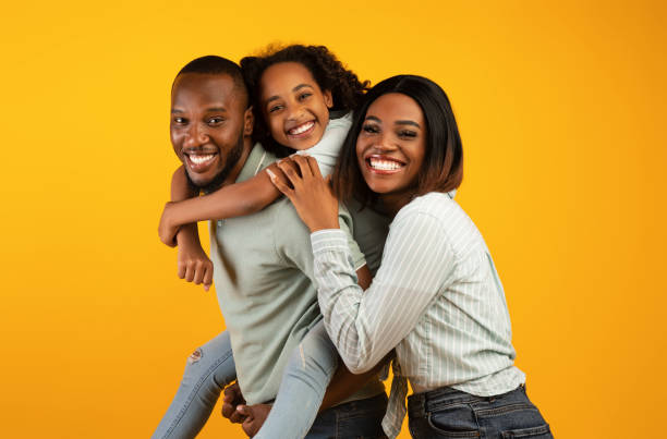 Time with family. Overjoyed african american family laughing and posing to camera isolated over yellow studio wall Time with family. Overjoyed african american family laughing and posing to camera isolated over yellow studio wall. Cheerful father carrying his daughter on back two parents photos stock pictures, royalty-free photos & images