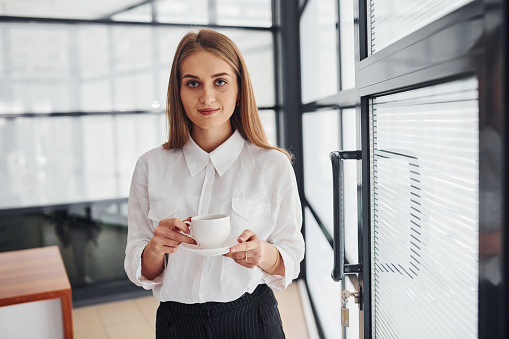Female office worker in formal clothes standing indoors at daytime with cup of drink.